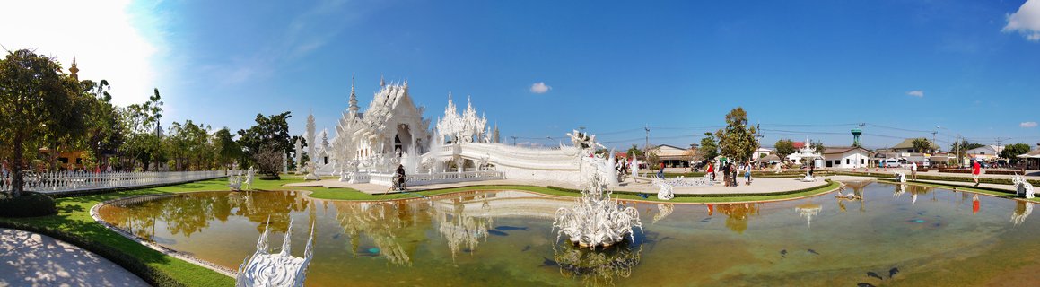 White ornate Buddhist temple complex with intricate spires and sculptures, reflection ponds in foreground, visitors walking on pathways, clear blue sky above, Chiang Rai Thailand
