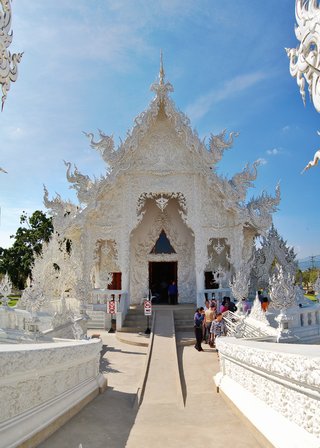 White ornate Thai Buddhist temple with elaborate carved decorations, naga serpent sculptures, and pointed gable roof against blue sky with tourists at entrance