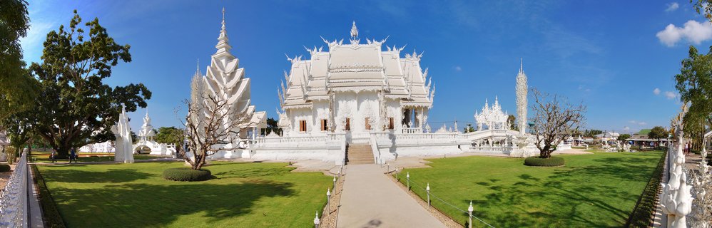White ornate Buddhist temple with multiple tiered roofs and intricate carvings surrounded by green lawns under blue sky