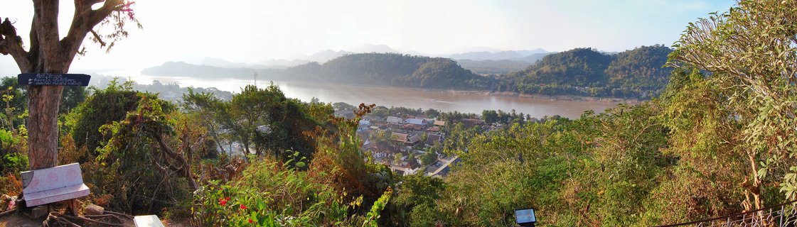 Wide panoramic view from elevated viewpoint showing Mekong River valley with forested mountains, traditional rooftops of Luang Prabang town, and tropical vegetation in foreground