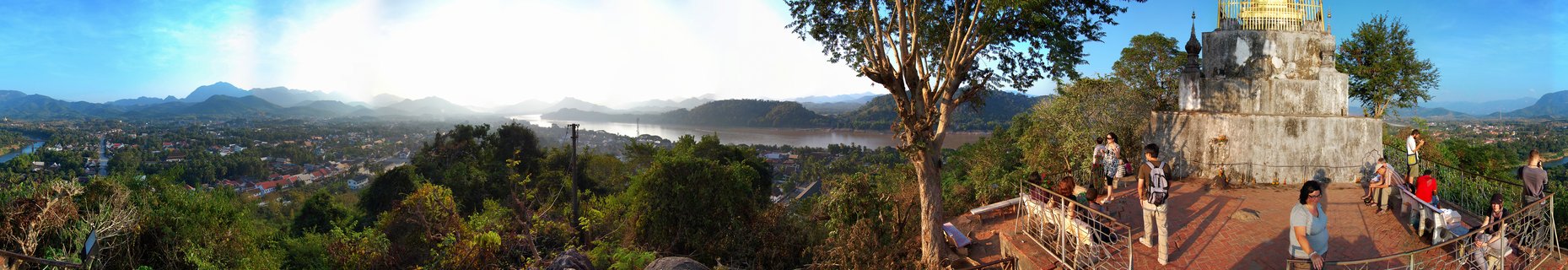Panoramic view from Mount Phousi showing Wat Chom Si temple with golden stupa, tourists on viewing platform, Luang Prabang city, Mekong River, and mountains in Laos