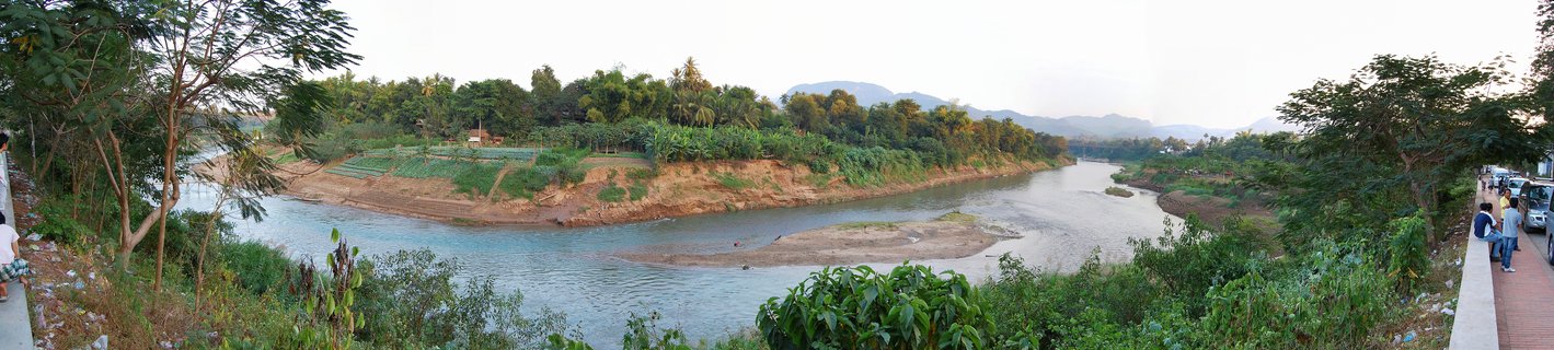 Wide panoramic landscape showing a river with sandbars flowing through green tropical vegetation, with mountains visible in the distance and people observing from a viewing platform on the right side