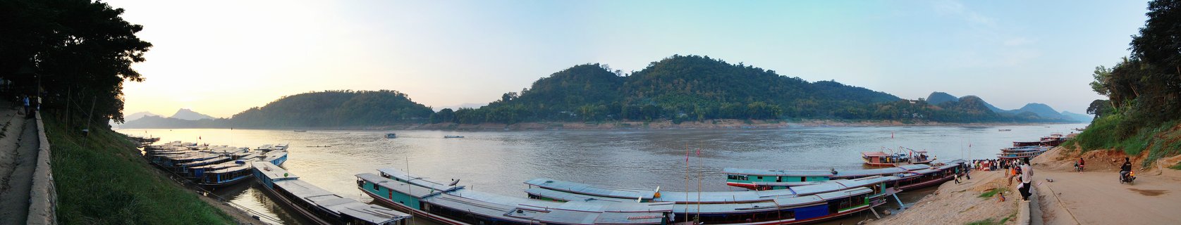Panoramic view of dozens of long wooden boats docked along the Mekong River shoreline with green hills in the background and people walking on the riverbank during late afternoon