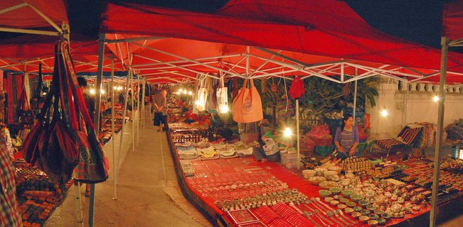 Night market stalls under red canopies with vendors selling handicrafts, textiles, ceramic bowls and jewelry on tables illuminated by hanging light bulbs