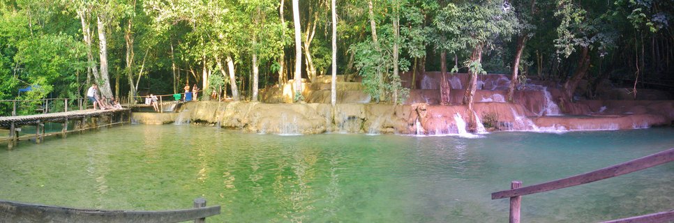 Multi-tiered waterfall with turquoise water flowing over limestone terraces, surrounded by green tropical trees with wooden walkways and tourists visible on the left side