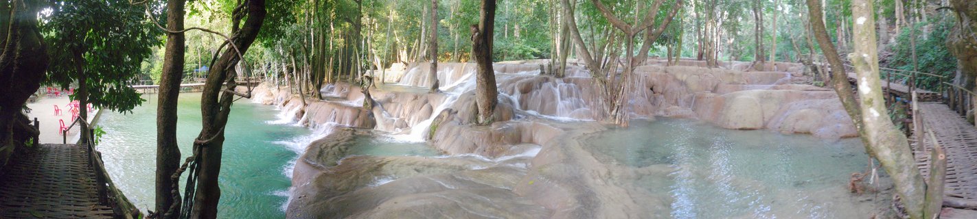 Multi-tiered waterfall with turquoise pools flowing over limestone terraces, surrounded by tropical forest with wooden walkways on both sides