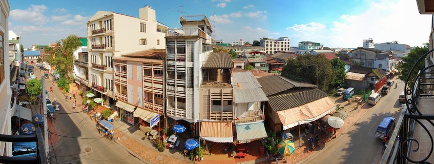 Panoramic street view of Vientiane, Laos showing buildings, shops with blue umbrellas, vehicles and pedestrians on intersecting roads under blue sky