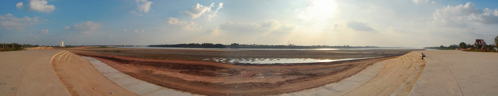 Panoramic view of concrete river embankments flanking an exposed muddy riverbed with water reflections, blue sky with clouds, Vientiane, Laos