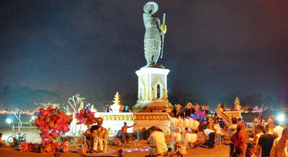 A large illuminated bronze statue of a standing figure holding a staff on an ornate white and gold pedestal at night, with crowds of people and street vendors selling colorful balloons and cotton candy at the base