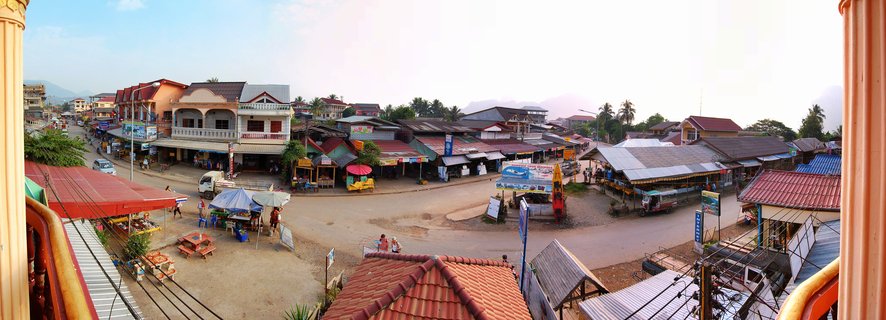 Panoramic street view of Vang Vieng town center showing market vendors with colorful umbrellas, buildings with red and metal roofs, people walking, and mountains in the background