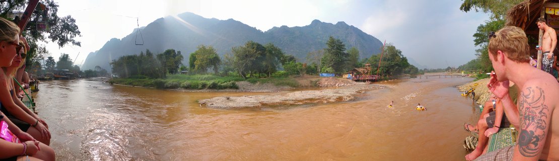 Wide panoramic view of tourists sitting at riverside bars and swimming in brown river water with large limestone mountains and tropical trees in background, Vang Vieng, Laos