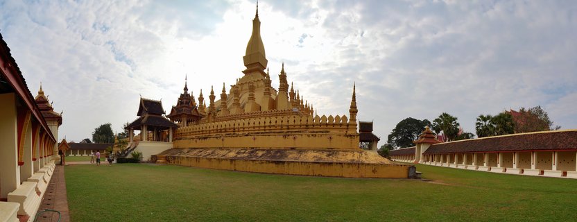 Golden Buddhist stupa with tall central spire surrounded by covered walkways and green lawn at Pha That Luang temple complex in Vientiane, Laos