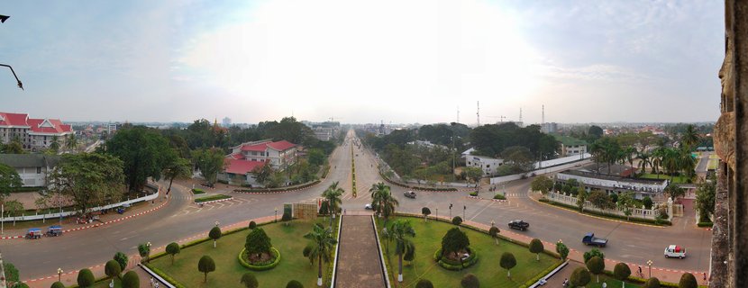 Panoramic aerial view of a wide tree-lined boulevard in Vientiane, Laos, with manicured gardens, government buildings with red roofs, traffic circles, and vehicles on the road under an overcast sky