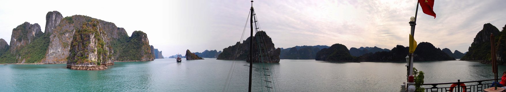 Panoramic view of limestone rock formations rising from calm turquoise water in Halong Bay, Vietnam, with boat deck and Vietnamese flag in foreground