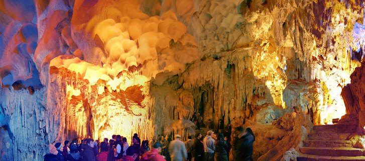 Interior view of illuminated limestone cave with hanging stalactites and rock formations, tourists walking below near stone stairs