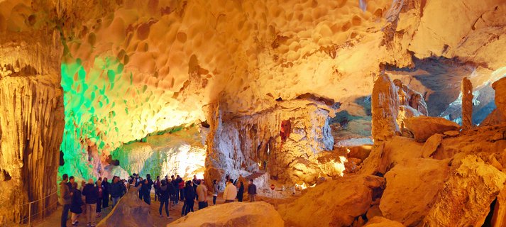 Interior view of a large limestone cave with tourists walking on a pathway. Stalactites hang from the ceiling and colored lights illuminate the rock formations in green and golden tones.