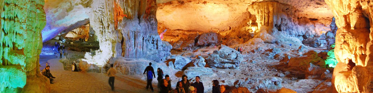 Interior view of a large limestone cave with tourists walking on pathways among illuminated stalactites and stalagmites with colored lighting in green, orange, and blue tones