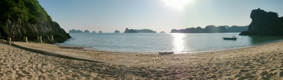 Wide beach scene showing sand in foreground, calm blue water with boats, and limestone rock formations and islands in the distance under bright sun at Halong Bay, Vietnam