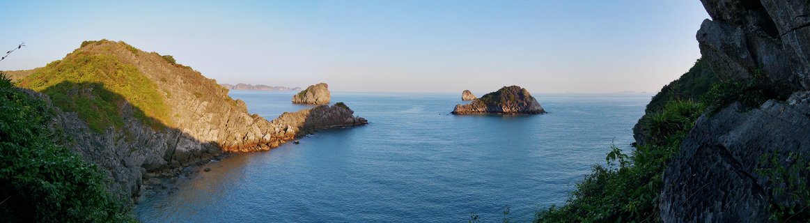 Rocky limestone islands covered with green vegetation rising from calm blue ocean water in Halong Bay, Vietnam with clear sky above