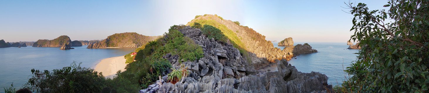 Panoramic seascape showing limestone karst islands covered in green vegetation rising from calm blue-green water in Halong Bay, Vietnam during late afternoon golden hour with rocky foreground and clear sky