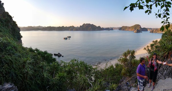 Panoramic view of Halong Bay showing limestone karst islands rising from calm water, with tourists at a clifftop viewpoint and cruise boats in the bay during golden hour