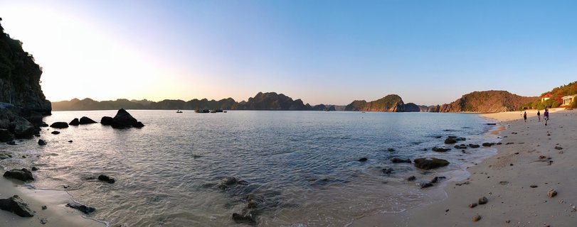 Panoramic coastal view showing limestone karst islands in calm bay waters with sandy beach, scattered rocks, and people walking along the shoreline under clear blue sky at golden hour