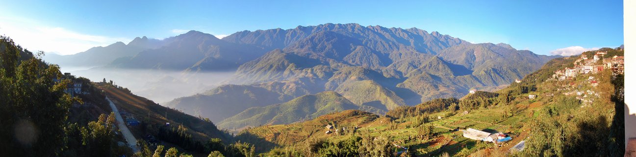 Panoramic mountain landscape showing terraced fields on green hillsides with valley fog and distant mountain ranges under clear blue sky in Sapa, Vietnam