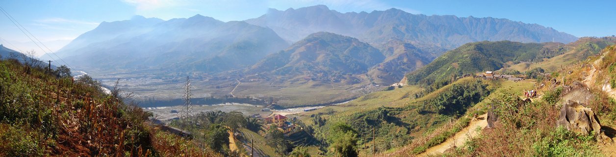 Wide mountain landscape showing green and brown hillsides with terraced fields, small buildings scattered on slopes, and multiple mountain ranges fading into blue haze in the distance under a clear sky