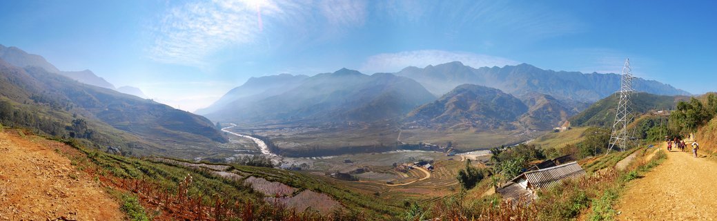 Panoramic landscape showing a mountain valley in Sapa, Vietnam with terraced rice fields on hillsides, a winding river below, distant mountains in morning mist, and a dirt path with people walking on the right side