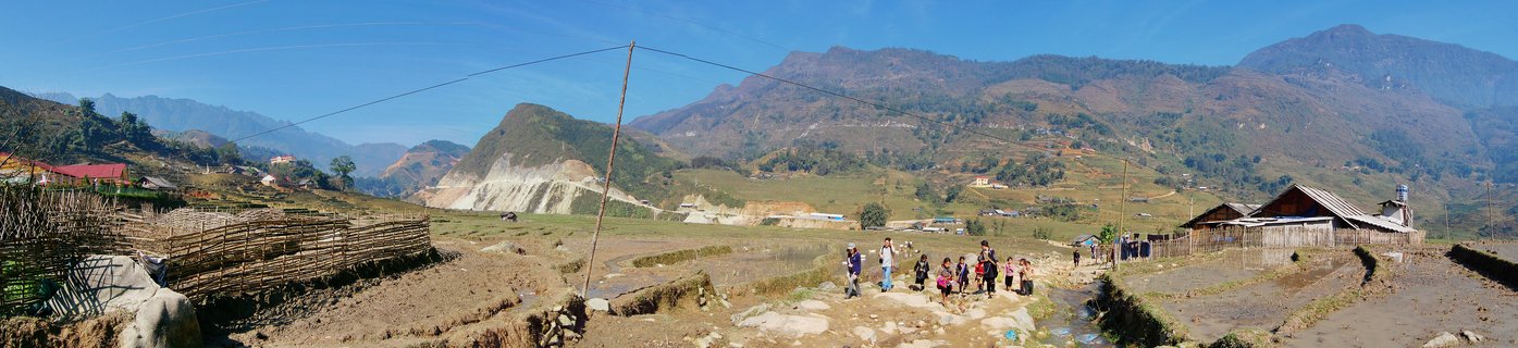 Panoramic view of rural village in Sapa Vietnam showing group of people walking on dirt path through dry terraced fields with traditional wooden houses and mountain range in background under clear blue sky