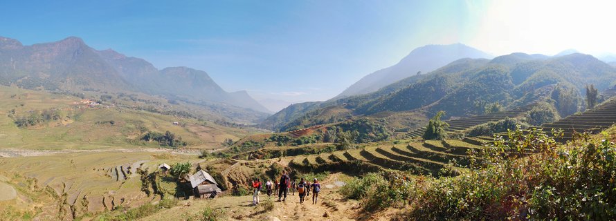 Group of hikers walking on dirt path through terraced rice fields with mountains in background under blue sky