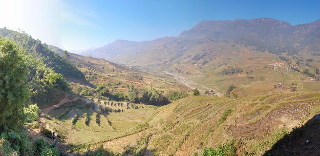 Wide panoramic landscape showing terraced rice fields on rolling hills in a valley with mountains in the background, clear blue sky, vegetation on hillsides, and a person walking on a path