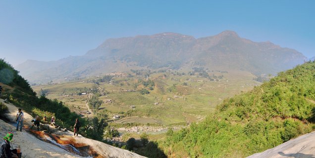 Panoramic landscape showing green terraced rice fields in a mountain valley with people in traditional clothing on rocks in the foreground and large mountains in the background under blue sky