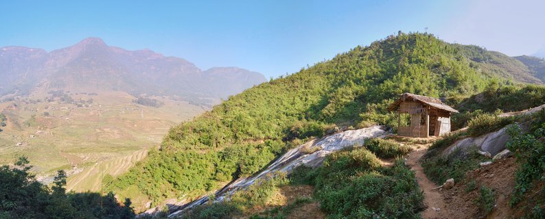 A small wooden hut on a green mountainside with terraced rice fields and distant mountains visible in the background under a clear blue sky in Sapa, Vietnam
