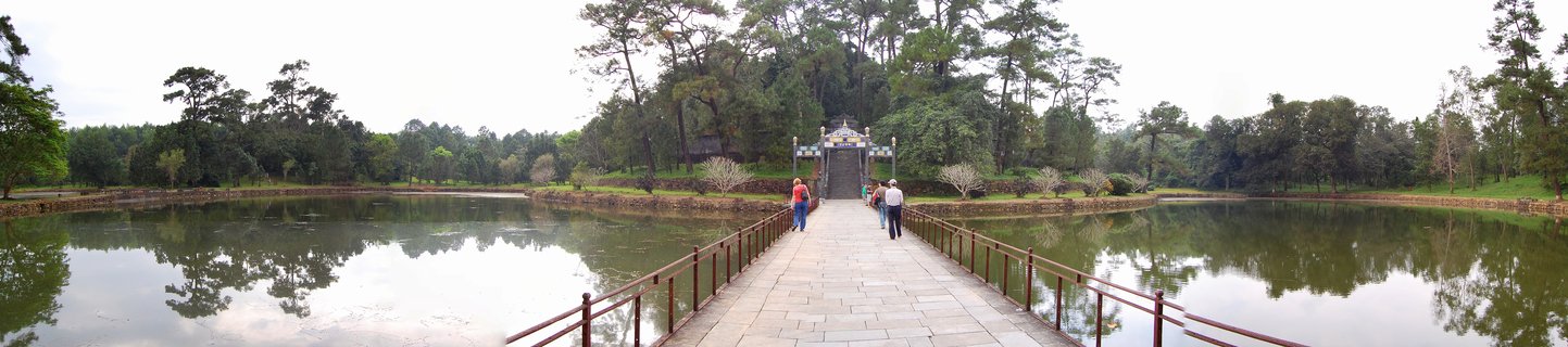 People walking on a long stone bridge with railings over calm water toward a traditional Vietnamese ornate gate surrounded by trees