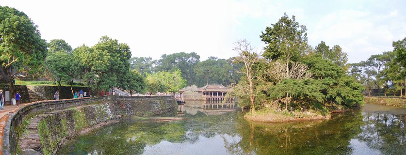 Wide view of Hue Imperial City showing traditional Vietnamese pavilion building across a moat with stone walls, trees, and tourists walking on pathway