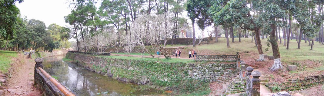 A canal with stone walls running through a park-like area with trees and people walking on pathways at Hue Imperial City in Vietnam
