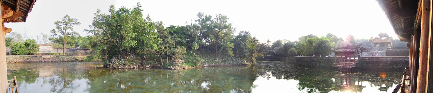 Wide panoramic view of a historic moat with traditional Vietnamese wooden pavilion, stone walls, tree-covered island, and water reflections at Hue Imperial City
