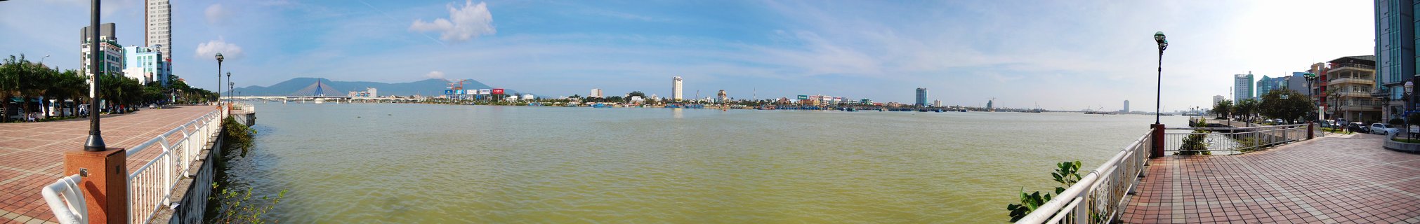 Wide panoramic photograph showing a river with paved walkways on both sides, city buildings along the banks, a bridge in the distance, and mountains on the horizon under a blue sky with scattered clouds.