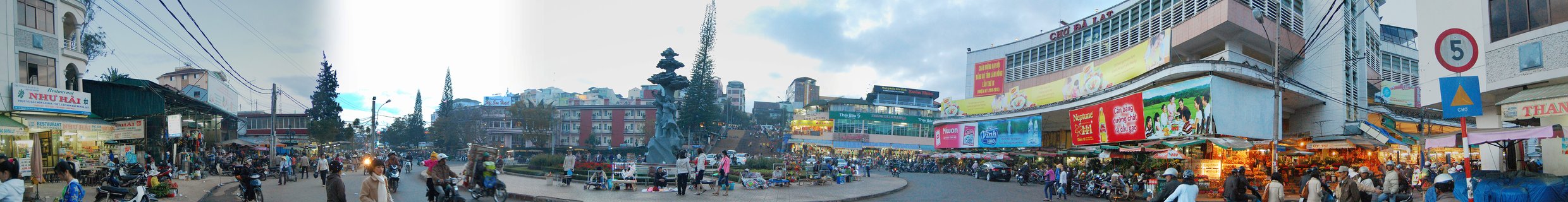 Panoramic street scene of Dalat city center square with central fountain, motorbikes, pedestrians, and market buildings under cloudy sky