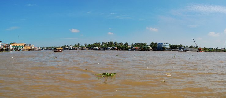 Wide river scene showing muddy brown Mekong Delta waters with boats, colorful riverside buildings on stilts, palm trees, and blue sky with scattered clouds