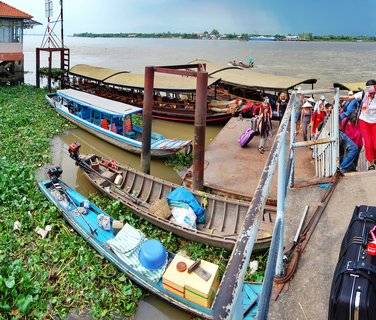A river port scene in Vietnam's Mekong Delta showing tourists with luggage on a floating dock, traditional covered boats with thatched roofs, small wooden sampans, and green water hyacinths covering parts of the brown river water
