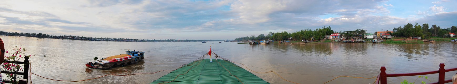 Wide river view from a green metal pier showing a cargo barge on brown water, with riverside buildings and tropical trees along the distant shore under cloudy sky