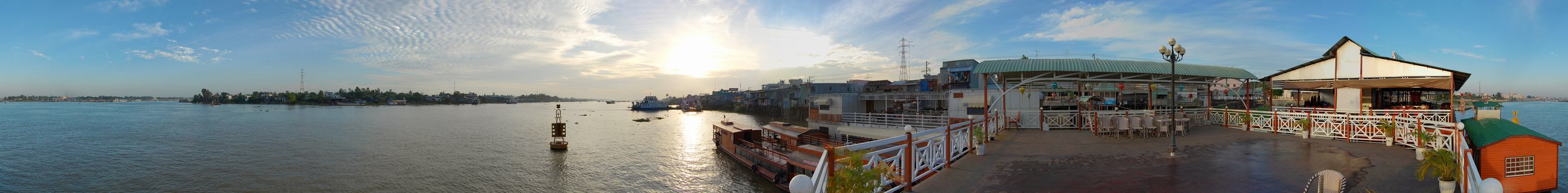 Panoramic photograph of a riverside restaurant terrace on the Mekong River in Phnom Penh at sunrise, with boats on the water and buildings along the waterfront under a blue sky with clouds
