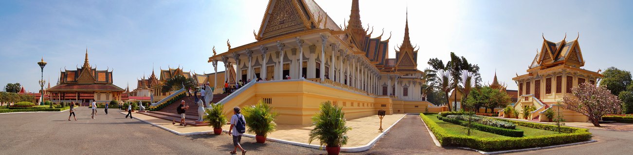 Wide view of the Royal Palace in Phnom Penh showing traditional Khmer buildings with golden pointed roofs, white and yellow walls, tourists walking on paved courtyards, and manicured gardens under clear blue sky