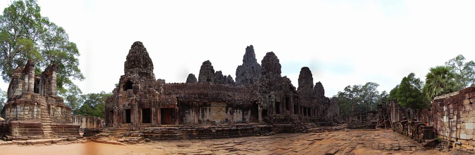 Panoramic view of ancient stone temple towers and ruins at Angkor Wat with tropical trees and stone courtyard under cloudy sky