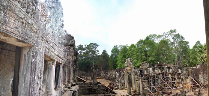 Ancient stone temple ruins with carved faces and columns at Bayon temple in Cambodia, with tropical trees in background and restoration scaffolding visible