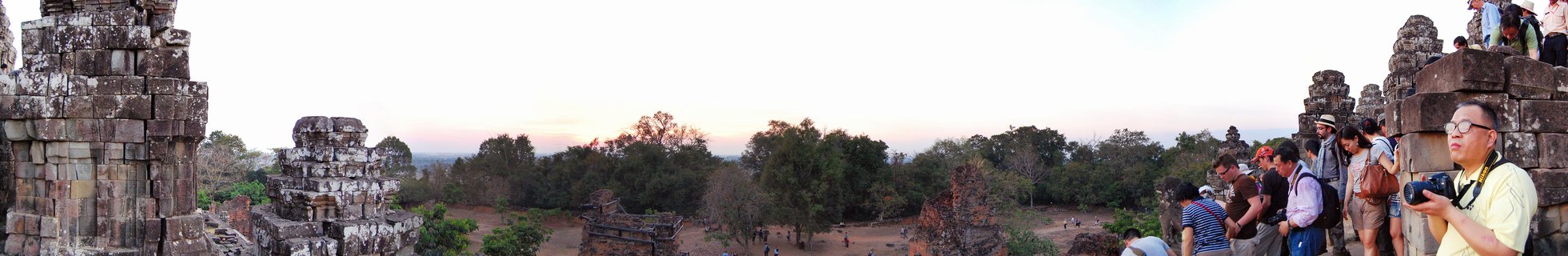 Panoramic view of ancient stone temple ruins at Angkor Wat with tourists standing on weathered sandstone structures, trees in the middle ground, and sunset sky above
