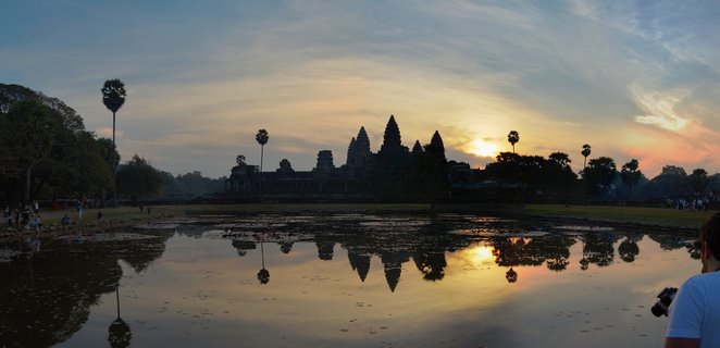 Angkor Wat temple complex at sunrise with five towers silhouetted against golden sky, reflected in foreground water with lotus pads and palm trees visible