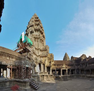 Stone temple tower of Angkor Wat with detailed carvings, scaffolding with green tarp on left side, stone courtyard and gallery corridors, clear blue sky above
